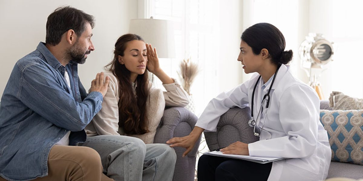 Serious young Indian female doctor telling about healthcare bad problems to couple of patients, talking to stressed shocked woman. Husband giving support to wife, listening to practitioner