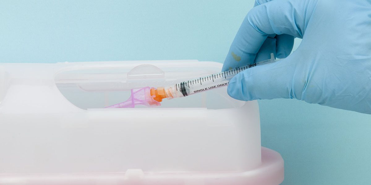 "This image shows a surgically gloved hand disposing of a syringe and needle(with safety cover) being disposed of in a safe,protective container known as a Sharp's container."